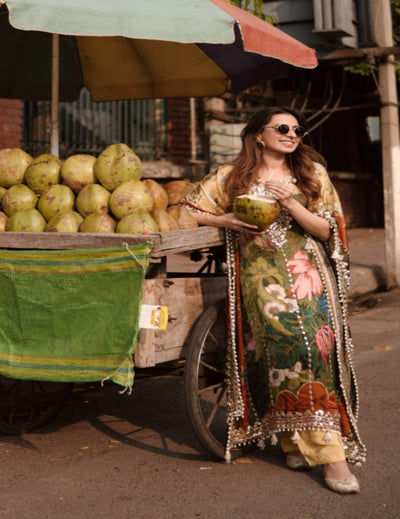 Woman in a colorful traditional outfit holding a coconut near a fruit cart.